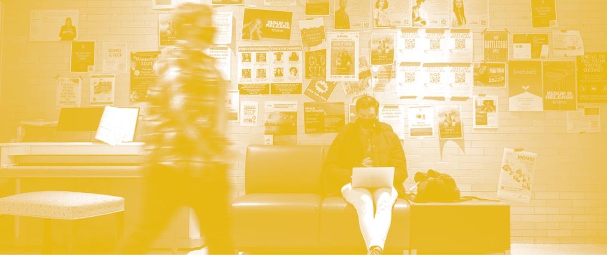 Yellow-tinted photo of a seated person in a mask working on a laptop in front of a crowded bulletin board