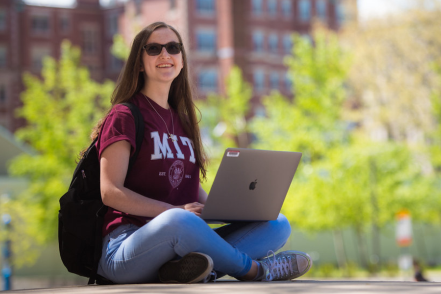 Student in a red MIT t-shirt sitting cross-legged outside with a laptop